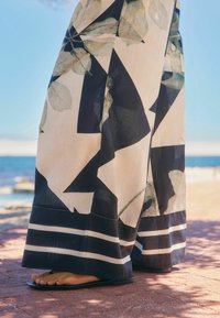 Gros plan sur une personne portant un pantalon large et fluide avec des motifs abstraits en noir et blanc et des feuilles, se tenant sur une surface en briques rouges près de la mer.