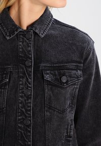 Giacca di jeans nera con colletto classico, chiusura anteriore con bottoni e taschino sul petto con una leggera texture usurata.