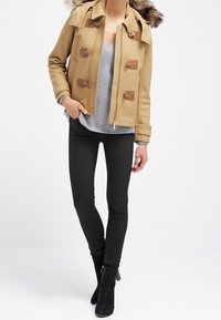 Vrouw in een camel kleurige dufflecoat met een bontkraag, grijze trui, zwarte skinny pants en zwarte enkellaarsjes die tegen een witte achtergrond staat.