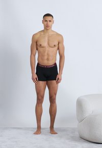 Μυώδης άντρας που στέκεται ξυπόλητος σε παρκέ δάπεδο, φορώντας μαύρα boxer briefs Tommy Hilfiger, δίπλα σε μια λευκή πολυτελή πολυθρόνα.