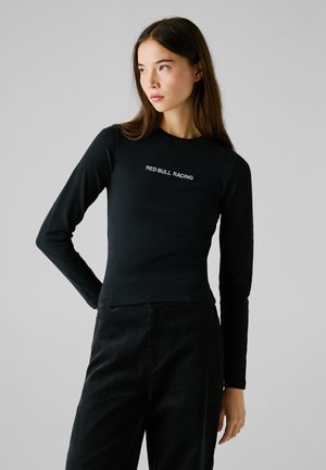 Young woman wearing a black long-sleeve shirt with "RED BULL RACING" text, paired with black pants, facing sideways against a gray background.