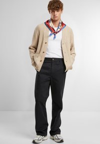 Beige ribbed cardigan with dark buttons over a white t-shirt, paired with dark pants and a colorful patterned scarf. Sneakers with mixed colors.