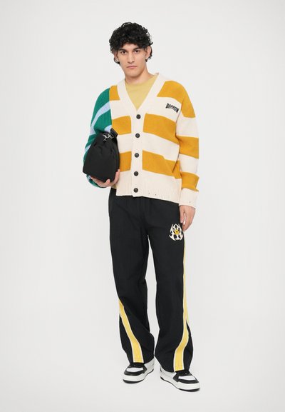 Striped cardigan w kolorze kremowym, żółtym i turkusowym; czarne spodnie z żółtymi paskami i logo; trzyma małą czarną torbę; wygodne obuwie.