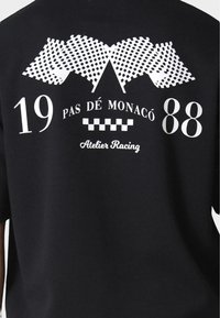 Fekete pamut póló, fehér grafikai dizájnnal, versenyszámokkal, "19 PAS DÉ MONACO 88" felirattal és "Atelier Racing" szöveggel.