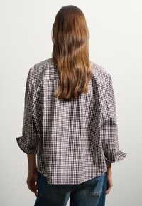 Persona con cabello largo castaño, vistiendo una camisa de cuadros marrón y blanca holgada y jeans azules, frente a una pared blanca sencilla.