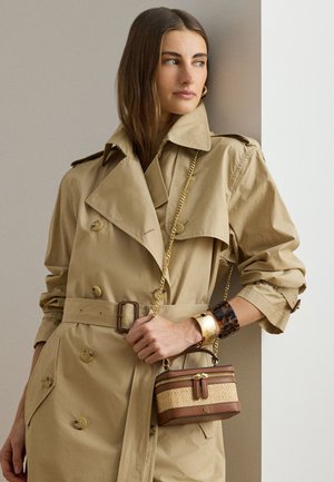 Γυναίκα με μπεζ καμπαρντίνα με μανίκια που έχουν γυριστεί, κρατά μικρή καφέ τσάντα με χρυσή αλυσίδα, φορώντας χρυσά και καφετιά βραχιόλια με σχέδιο χελώνας.