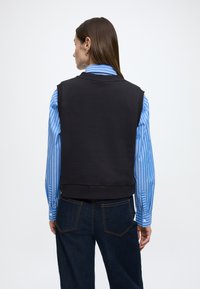 Colete preto sem mangas com textura suave, usado sobre uma camisa às riscas azuis e brancas. Apresenta um decote arredondado e bainha canelada.