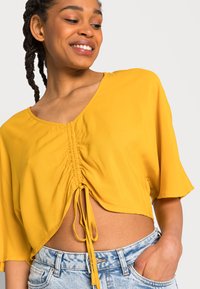 Top cropped amarelo de manga curta com decote em V e centro franzido com detalhes em laço. Combinado com calções de ganga azul claro.