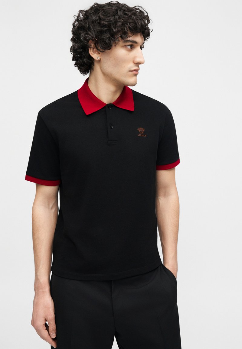 Man wearing black polo shirt with red collar and sleeve edges, looking to the right, hands in pockets, standing against a white background.