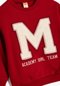 Felpa rossa con un grande logo "M" in chenille bianco, con contorni ricamati e il testo "ACADEMY GIRL TEAM" sotto. Tessuto testurizzato.