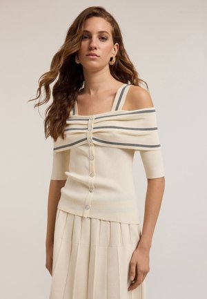 Beige ribbad cardigan som går av axlarna med korta ärmar, central knappstängning och blå randiga detaljer. Slät textur och figurnära design.