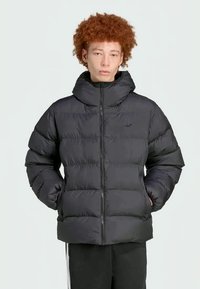 Mlad človek s mahu rdečimi lasmi, oblečen v črn puhasti plašč, z rokami v žepih, stoji pred enostavno svetlo ozadje.