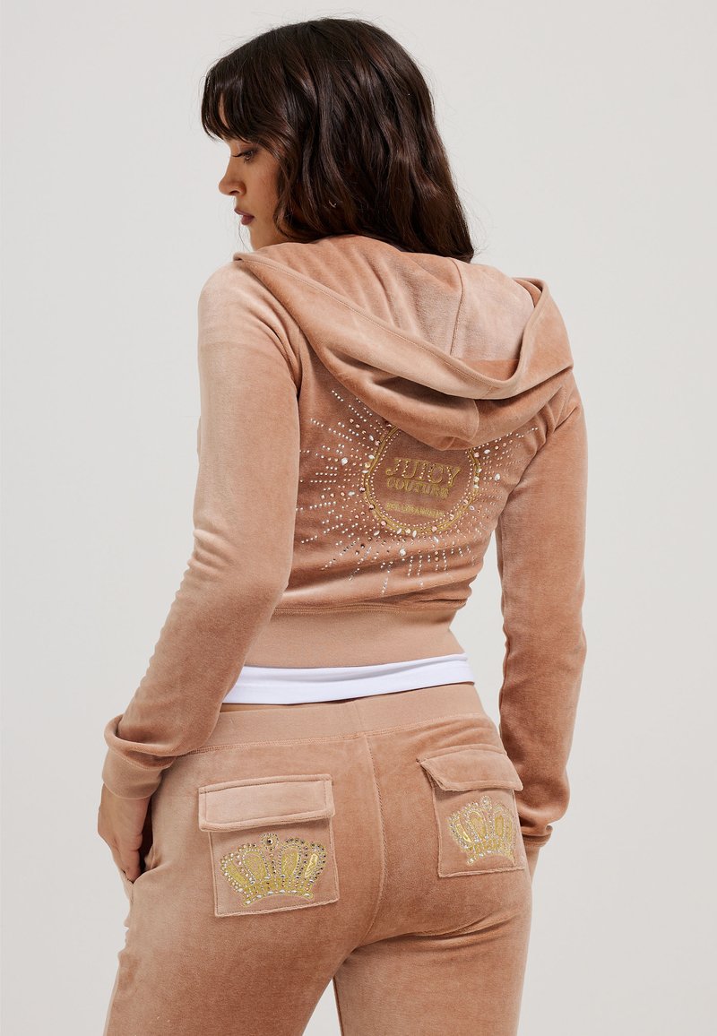 Tuta in velluto rosa con cappuccio. Il retro presenta un logo dorato e dettagli in strass. I pantaloni hanno disegni di corone sulle tasche.