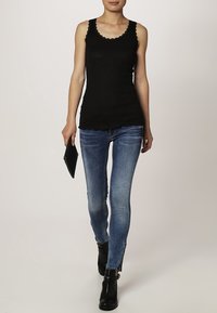 Zwarte tanktop met geschulpte halslijn, gedragen met lichtblauwe skinny jeans en zwarte enkellaarsjes, met een zwarte clutch. Effen achtergrond.