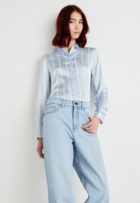 Camisa de botão em satin azul claro com riscas bejes verticais, gola clássica e mangas longas, combinada com jeans azul claro de cintura alta.