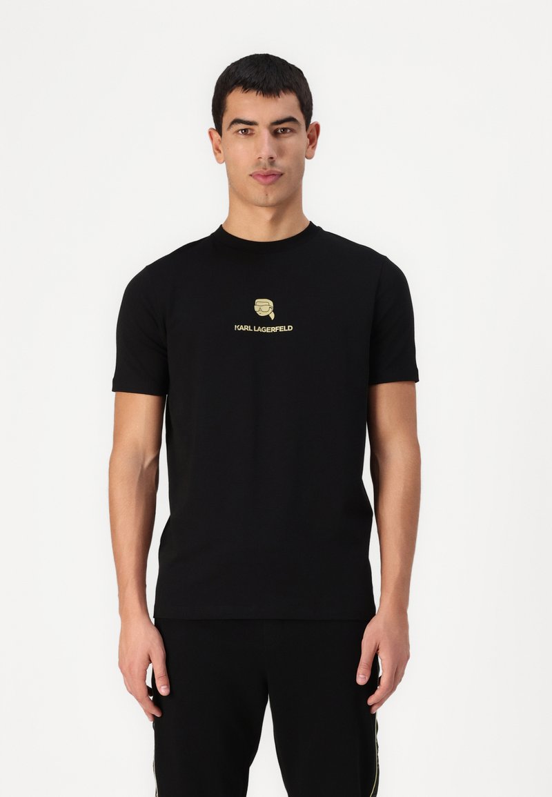 Juodas medvilnės marškinėliai su apvalia kaklo iškirpte, trumpomis rankovėmis ir auksiniu logotipu, vaizduojančiu stilizuotą galvą su užrašu "KARL LAGERFELD".