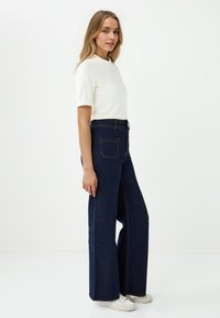 Jeans in denim scuro a gamba larga con cuciture arancioni a contrasto, caratterizzati da un vita alta, due tasche posteriori e una texture liscia. Abbinati a una maglietta bianca.