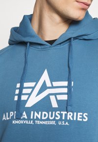 Blå bomullshuvtröja med en stor vit logotyp med geometriska mönster och texten "ALPHA INDUSTRIES, KNOXVILLE, TENNESSEE, U.S.A."