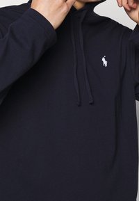 Navy modrá bavlněná mikina s kapucí, klokanní kapsou a malým vyšívaným bílým logem na levé hrudi.