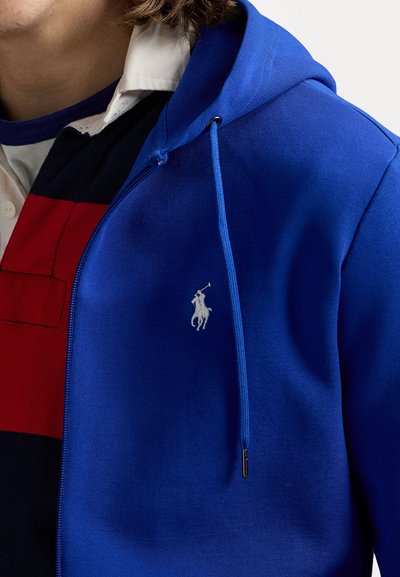 Pessoa vestindo uma sweatshirt azul com fecho éclair e cordões, por cima de uma camisola de rugby às riscas em vermelho, preto e branco.