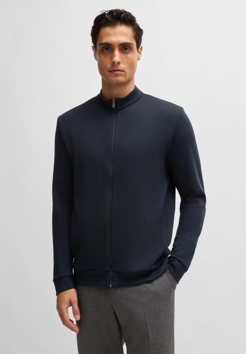 Veste zippée marine à surface texturée, col montant et poignets côtelés. Portée sur un pantalon gris, avec un design épuré.