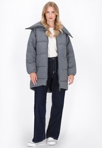 Grå pufferjakke med bred krave, vandret syninger, lynlås foran og sidelommer, parret med mørke jeans og hvide sneakers.
