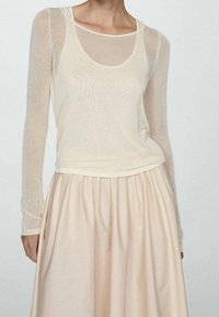 Top ligero y translúcido de manga larga en crema, superpuesto a una camiseta de escote redondo, combinado con una falda fluida de color rosa pálido con cintura fruncida.