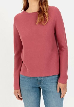 Femme portant un pull rose à manches longues et col rond ainsi qu'un jean bleu clair, debout devant un fond blanc uni.