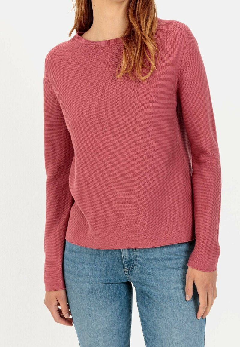 Femme portant un pull rose à manches longues et col rond ainsi qu'un jean bleu clair, debout devant un fond blanc uni.