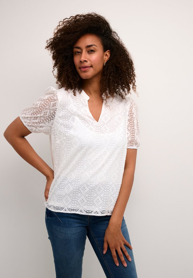 Blouse blanche avec un design en dentelle géométrique, manches courtes bouffantes et décolleté en V. Portée avec un jean bleu. Détails de tissu texturé visibles.
