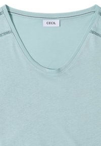 Haut bleu clair à col rond avec une délicate bordure en dentelle sur les épaules et une petite étiquette rectangulaire portant l’inscription "CECIL" à l’encolure.