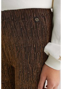 Juodos ir bronzinės spalvos raštuotas audinys su vertikaliomis juostelėmis ant kelnių, kurių juosmens dalyje yra auksinė logotipo detalė, derinama su balta rankovėmis.