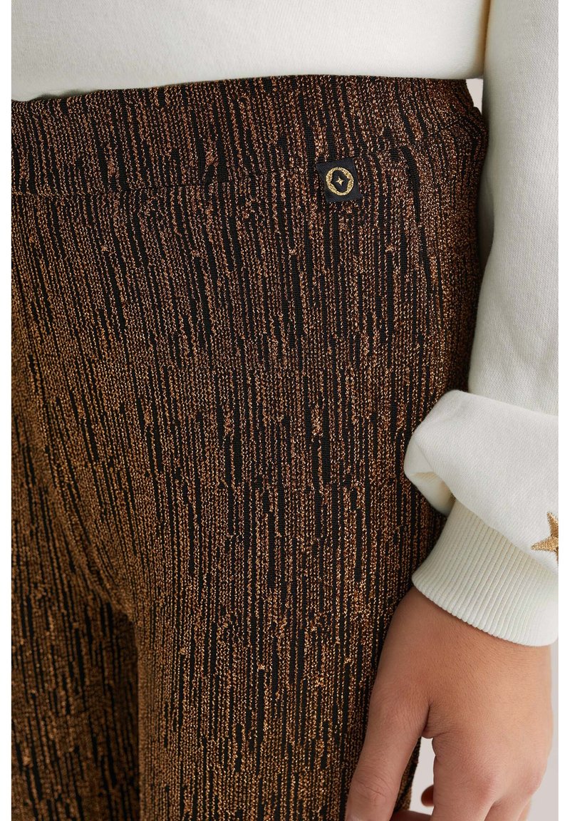 Juodos ir bronzinės spalvos raštuotas audinys su vertikaliomis juostelėmis ant kelnių, kurių juosmens dalyje yra auksinė logotipo detalė, derinama su balta rankovėmis.