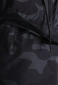 Material negru cu un model subtil de camuflaj, având o buzunar cu fermoar și accente cu șnur. Textură netedă și design ușor.