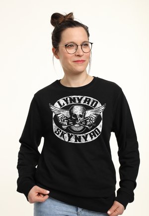 Kvinde med briller og hår i en knold iført en sort Lynyrd Skynyrd sweatshirt og lyseblå jeans mod en hvid baggrund.