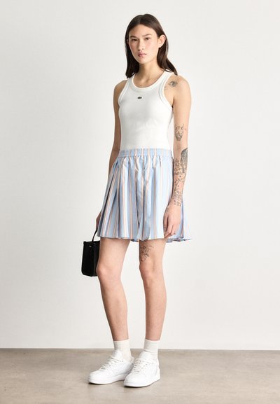 Débardeur blanc avec petit logo, jupe plissée bleue à rayures, baskets blanches et sac à main noir. Tissu lisse, design décontracté.