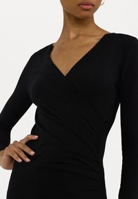 Mujer con vestido envolvente negro de manga larga y escote en V, con la mano en la cadera, contra un fondo blanco liso.