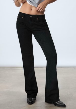 Osoba ubrana w czarne jeansy bootcut i czarne szpiczaste buty, podnosząca szarą koszulę, aby odsłonić brzuch na jednolitym tle.