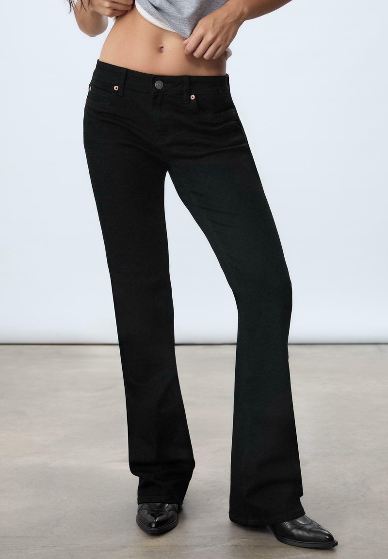 Personne portant un jean bootcut noir et des chaussures pointues noires, soulevant une chemise grise pour révéler le ventre sur un fond uni.