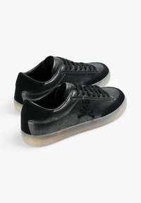 Pareja de zapatillas negras de corte bajo con textura brillante, detalles de ante, cordones y suelas de goma translúcidas sobre un fondo blanco.