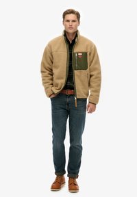 Beżowa kurtka polarowa z zieloną kieszenią na piersi, zapinana na suwak, z kontrastowymi brązowymi skórzanymi elementami; w zestawie z niebieskimi jeansami i brązowymi butami.