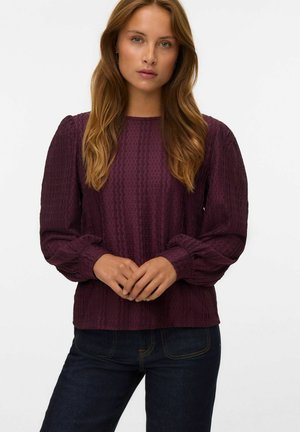 Blouse à manches longues, texturée, en bordeaux profond avec un motif côtelé. Présente un col rond et des poignets froncés, associée à un jean noir.