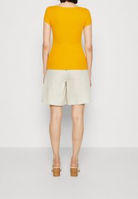 Gul, ribbad kortärmad topp i kombination med ljusbeige linnebyxor och sandaler med brun klack, sedda bakifrån.