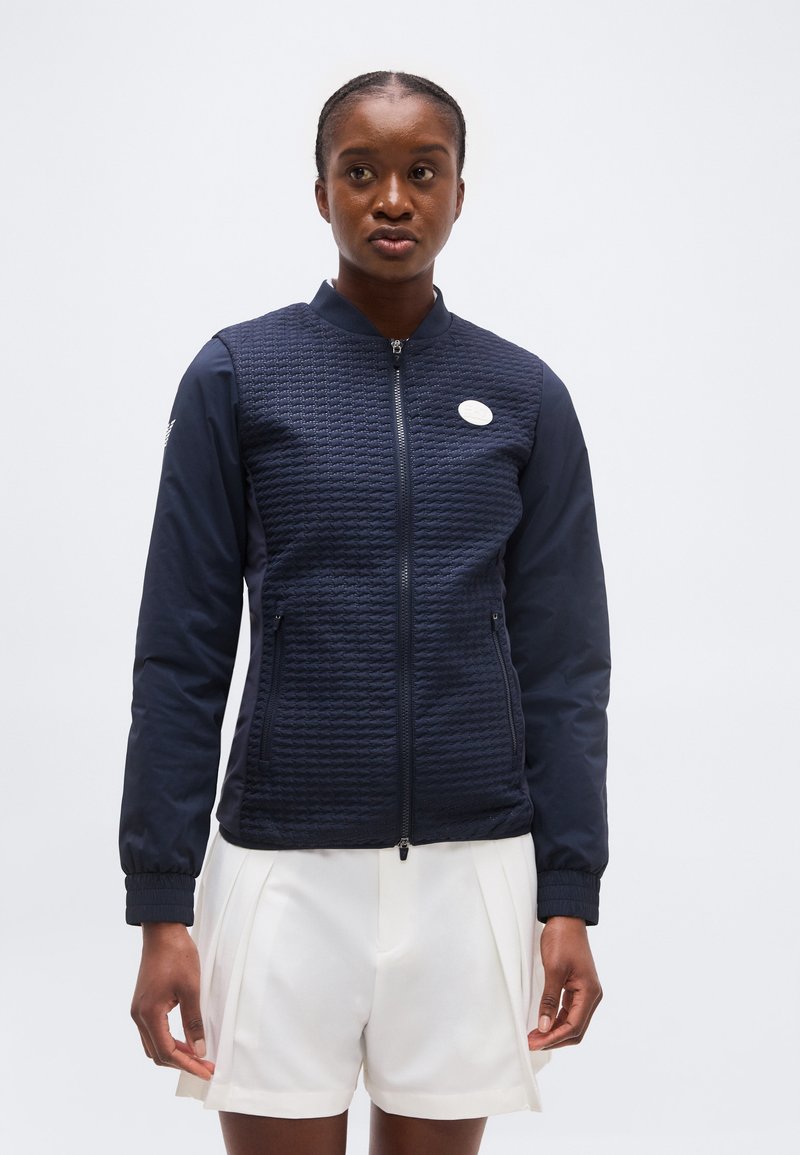 Γυναίκα που φοράει μπλε ναυτικό μπουφάν με φερμουάρ και ελαστικά μανίκια, συνδυασμένο με λευκό σορτς, στέκεται μπροστά σε απλό φωτεινό φόντο.