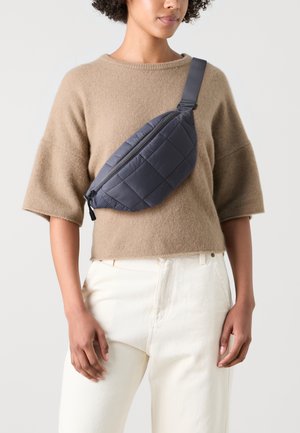 Quiltet grå midjeväska med dragkedjestängning, tillverkad av lätt material, bärs över kroppen, kombinerad med en beige tröja och vita byxor.