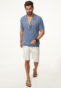 Blaues gestricktes Kurzarmhemd mit Knopfleiste, kombiniert mit weißen strukturierten Shorts und braunen Sandalen. Modell steht vor einem neutralen Hintergrund.