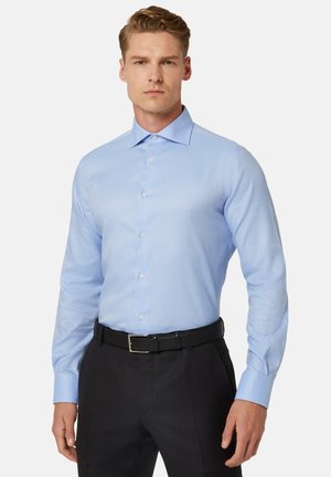 Camicia azzurra chiara con maniche lunghe, colletto a bottoni e patta frontale. Indossata con cintura nera e pantaloni scuri.