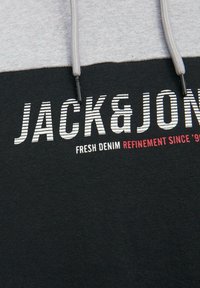Mikina s kapucňou so čiernym telom a šedými rukávmi, s šnúrkami a potlačou v bielej a červenej farbe, zdôrazňujúca značku a rafinovanosť.