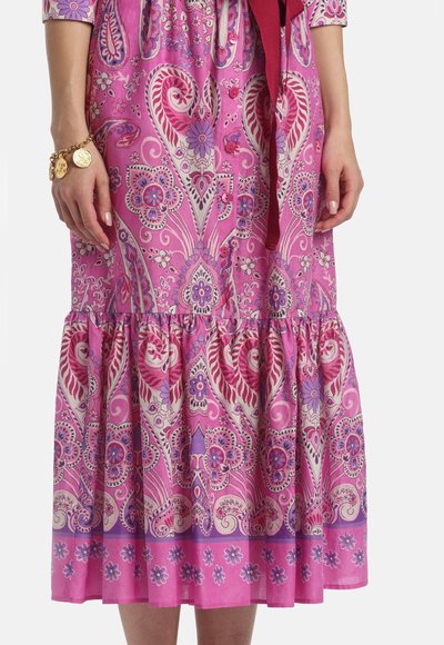 Kvinde iført en midi-kjole med pink og lilla blomster- og paisleymønster, knapper foran og en flæsekant, med viste hænder og armbånd.