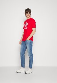 Camiseta de algodón roja con logo blanco de Adidas, vaqueros de mezclilla azules y zapatillas deportivas beige. Hombre con gafas de sol negras. Fondo simple.